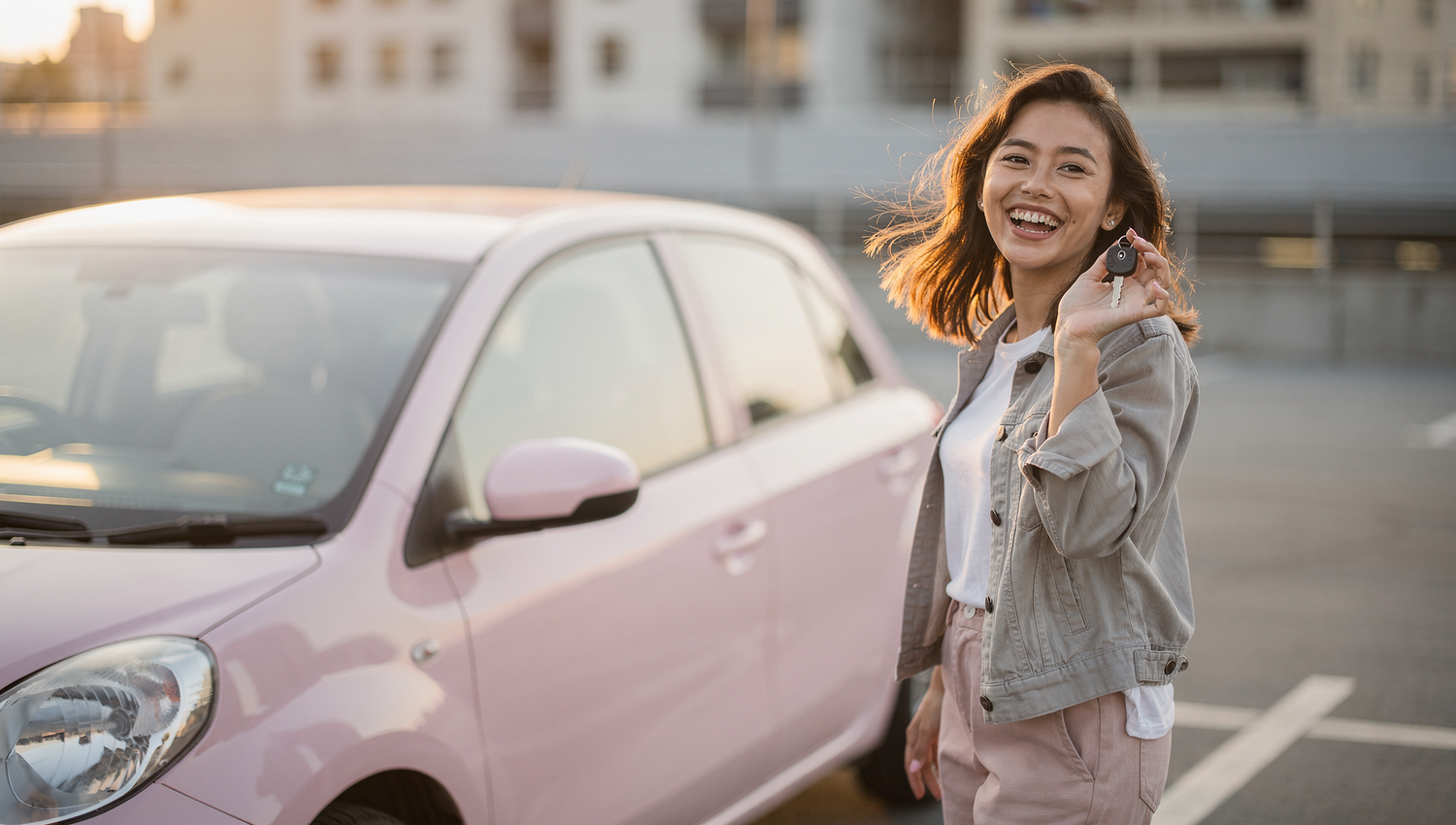 Confident car buying woman with keys after a successful purchase of a pink used car in a blurred urban setting